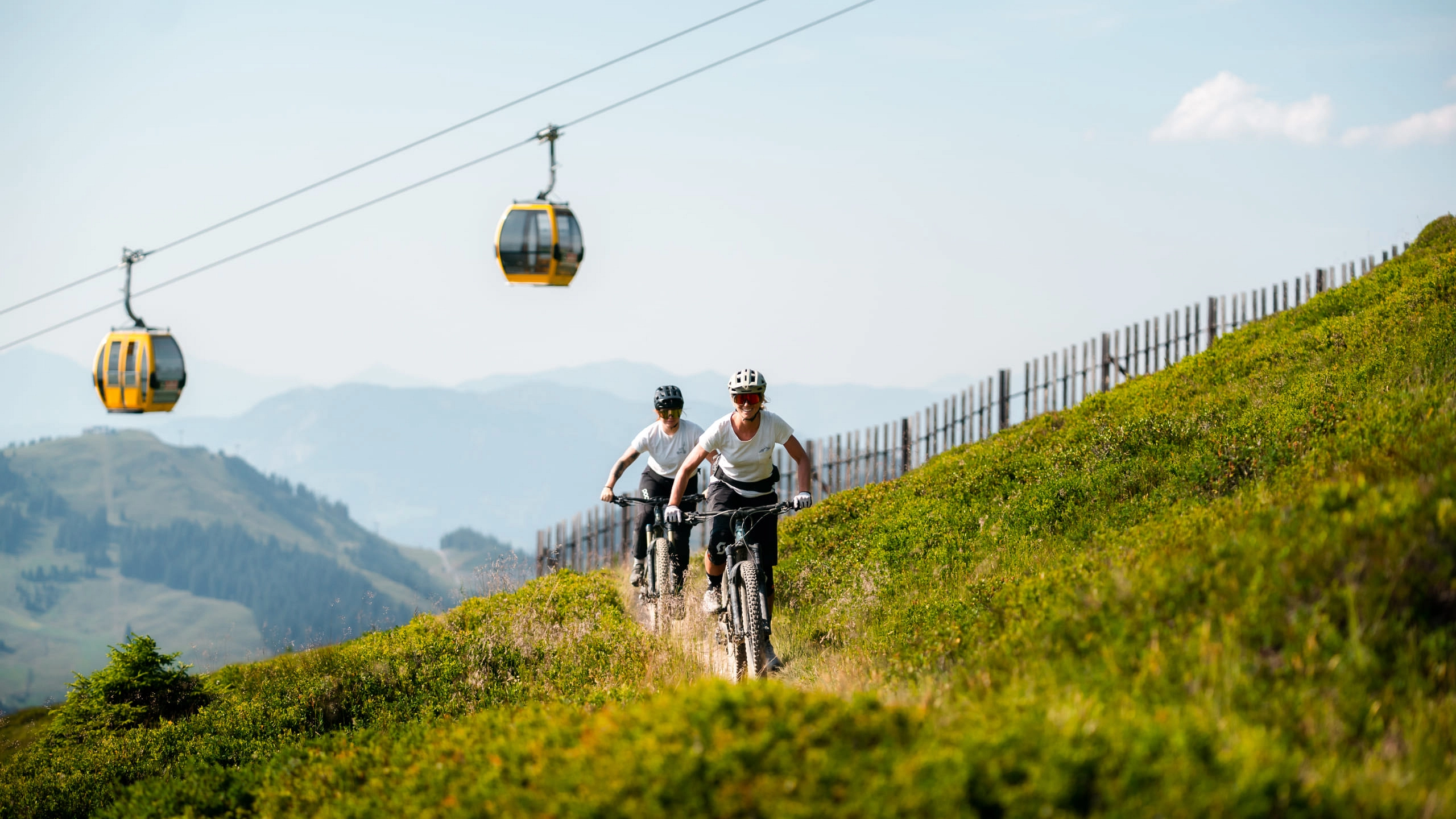 Zwei Radfahrer auf einem sonnigen Bergweg mit Seilbahnkabinen im Hintergrund