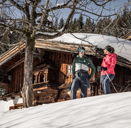 Zwei Personen wandern durch schneebedecktes Gelände vor einem Holzhaus.