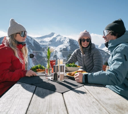 Drei Personen essen draußen mit Bergblick, Speisen und Getränke auf dem Tisch.